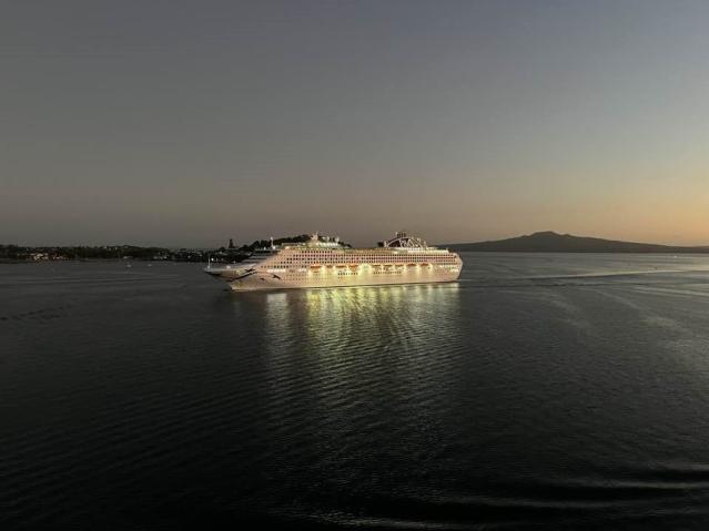 A illuminated cruise ship sails on calm waters at dusk. The ship's lights reflect off the dark sea, creating a glowing effect.